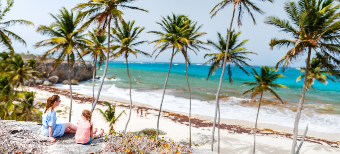 Zwei Frauen – eine Erwachsene und ein Kind – sitzen auf einem Felsen mit Blick auf einen palmengesäumten Strand und das türkisblaue Meer. Die Szene ist von oben aufgenommen und zeigt Palmen, Felsen, Wellen und eine naturbelassene Küstenlandschaft bei hell