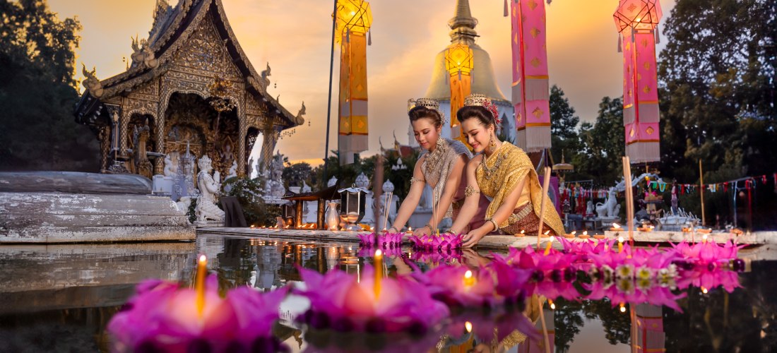 Das Bild zeigt zwei thailändische Frauen in prachtvoller traditioneller Kleidung, die an einem festlich beleuchteten Tempel in der Dämmerung schwimmende Lichter und Blütenkränze ins Wasser setzen – sogenannte Krathongs. Im Hintergrund steht ein kunstvoll 