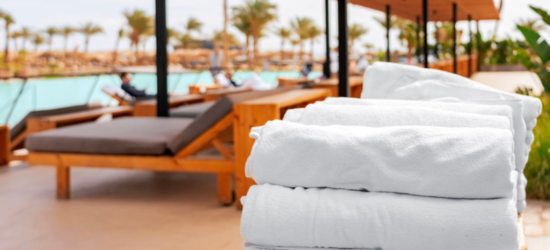 Gestapelte weiße Poolhandtücher im Vordergrund, dahinter stilvolle Holzliegen mit Auflagen unter einem schattenspendenden Dach am Pool eines Resorts. Im Hintergrund Palmen und entspannte Gäste.