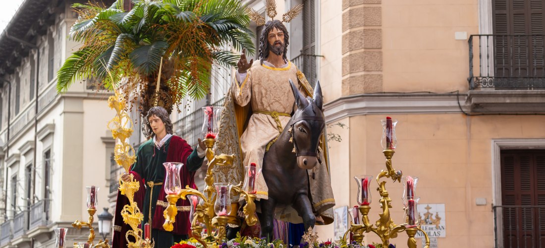 Eine festliche Prozession zeigt eine Figur von Jesus auf einem Esel, begleitet von einer weiteren Figur, auf einer prächtigen Plattform mit Palmen, Blumen und goldenen Kerzenhaltern. Die Jesusfigur trägt ein helles Gewand mit goldener Stickerei und hat go