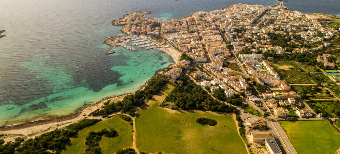 Luftaufnahme einer Küstenstadt auf Mallorca mit einem langen Sandstrand und türkisfarbenem Wasser. Im Vordergrund ist eine weitläufige grüne Landschaft mit Feldern und Bäumen zu sehen. Die Stadt liegt direkt am Wasser und hat einen kleinen Yachthafen. Das