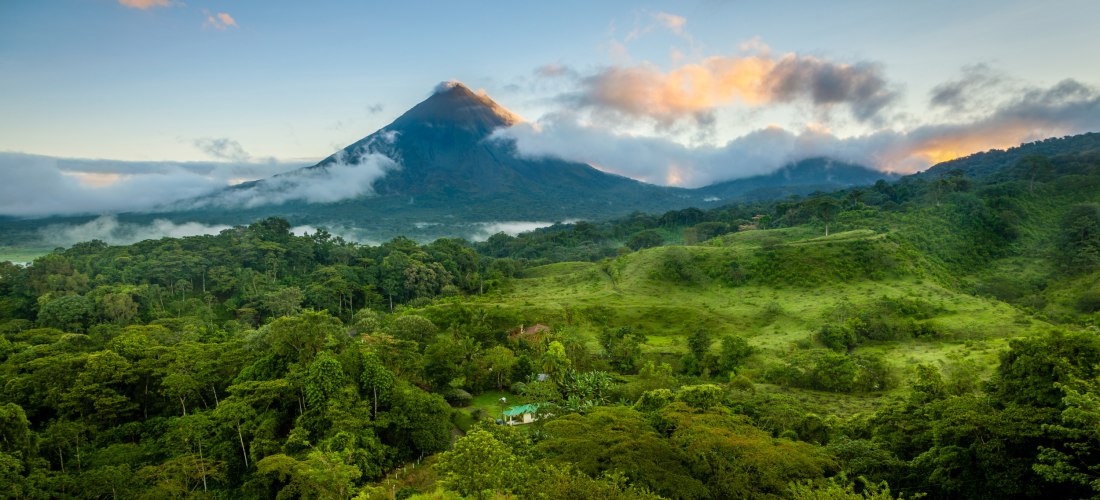 Üppig grüne tropische Landschaft mit dichtem Regenwald und dem Vulkan Arenal im Hintergrund, teilweise von Wolken umhüllt, bei Sonnenaufgang