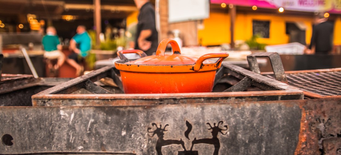 Ein oranger Gusseisentopf steht auf einem traditionellen Grill im Freien, eingerahmt von Metall mit geschnitzten Symbolen. Im Hintergrund Menschen und eine farbenfrohe Kulisse – eine karibische Straßenszene mit lebendiger Atmosphäre.