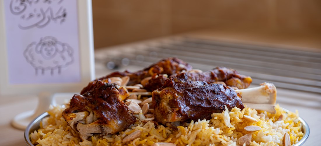 Ein traditionelles arabisches Reisgericht mit gewürztem Lammfleisch und Mandeln auf einem großen Metallteller. Daneben eine festliche Karte mit arabischer Kalligraphie und einer Zeichnung eines Schafs – typisch für das Opferfest (Eid al-Adha).