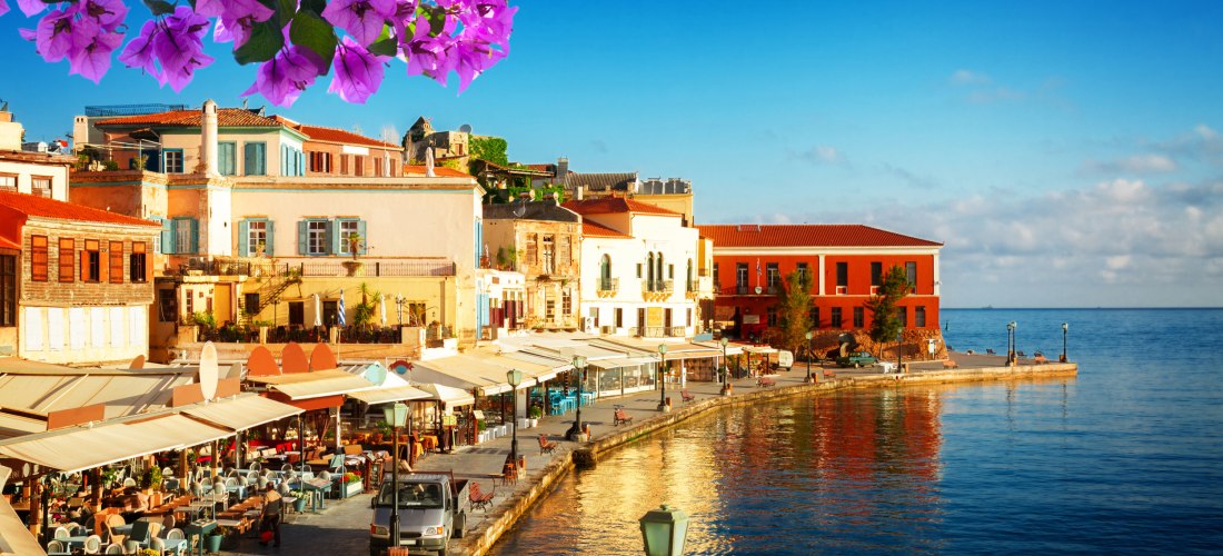 Hafenpromenade von Chania auf Kreta mit bunten Häusern, Straßencafés und lila Bougainvillea-Blüten unter blauem Himmel