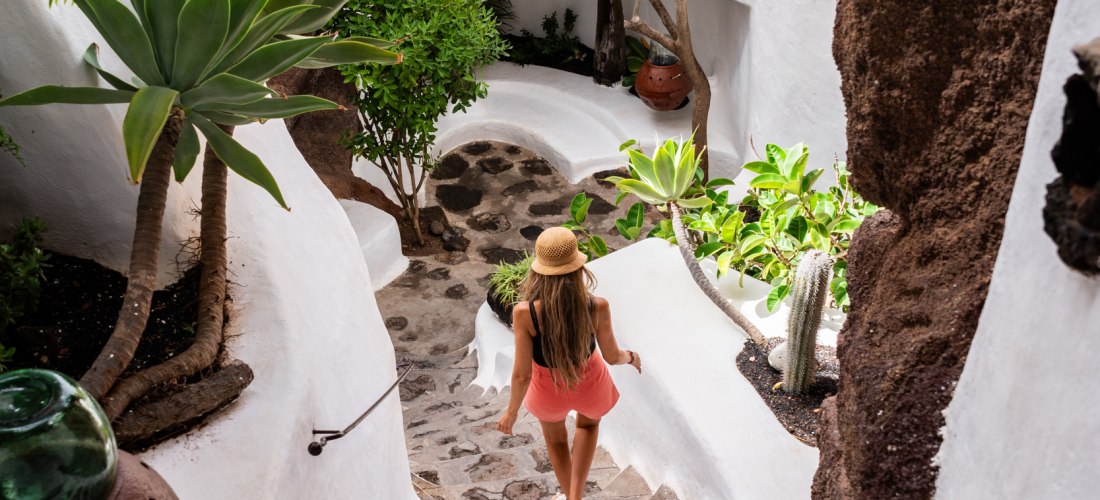 Frau mit Strohhut und Sommerkleid geht eine steinerne Treppe hinunter, umgeben von weiß verputzten Wänden, Pflanzen und Felsen in organisch geschwungener Architektur