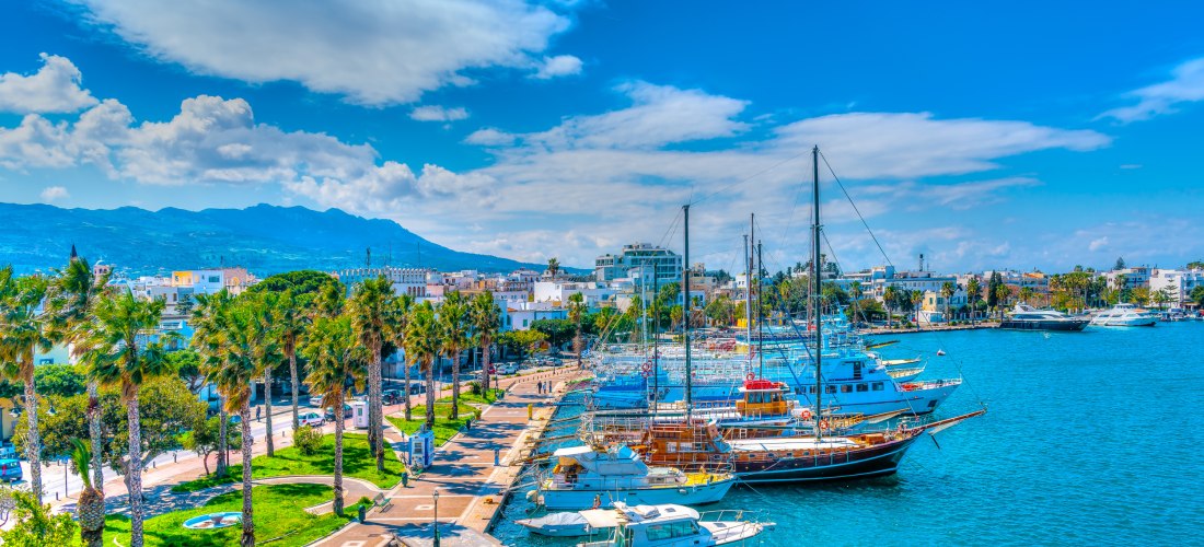 Panorama einer Hafenpromenade mit Palmen, gepflegten Wegen und zahlreichen Segel- und Motorbooten im türkisfarbenen Wasser. Im Hintergrund erstreckt sich eine Stadt mit weißen Häusern, darüber eine imposante Bergkette unter blauem Himmel mit dekorativen W