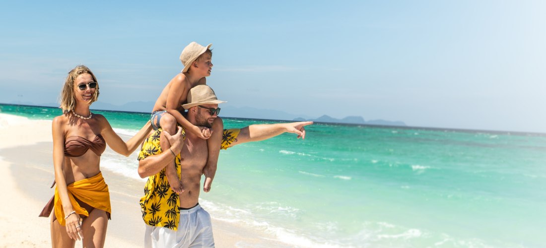 Eine fröhliche Familie spaziert am Strand entlang. Der Vater trägt das Kind auf den Schultern und zeigt lachend aufs Meer, während die Mutter daneben geht. Alle tragen Sommerkleidung. Türkisfarbenes Wasser und blauer Himmel im Hintergrund