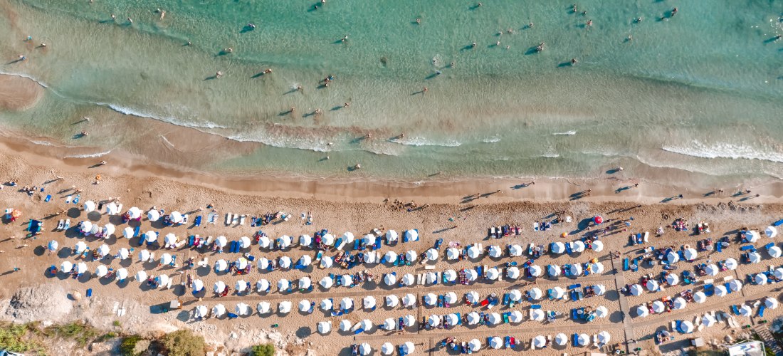 Drohnenaufnahme eines belebten Sandstrands auf Zypern mit Reihen weißer Sonnenschirme und Badegästen im flachen, türkisfarbenen Wasser.