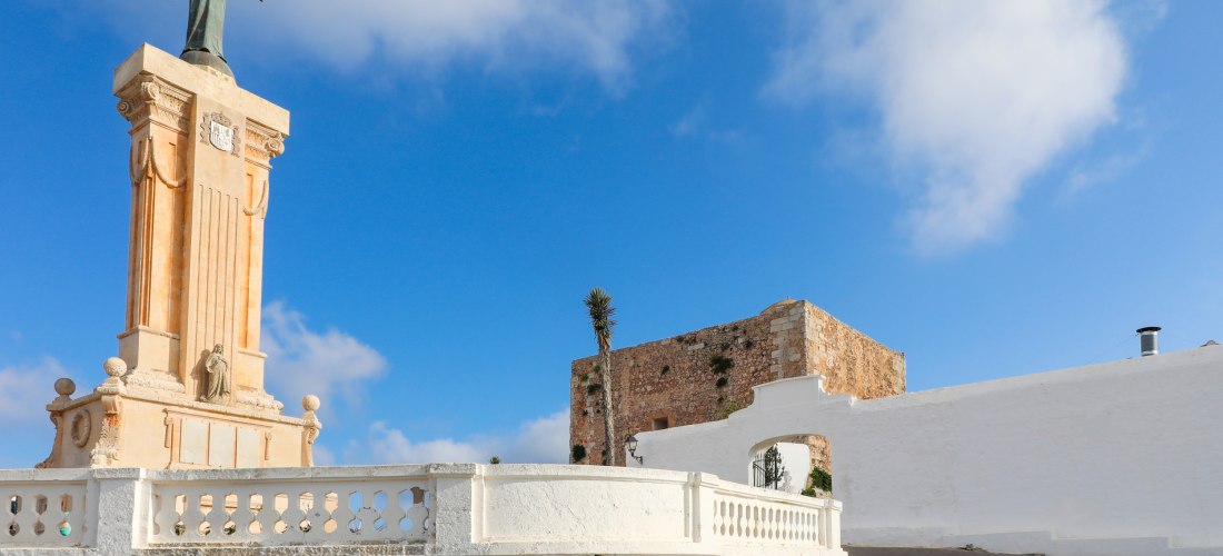 Statue von Christus mit ausgebreiteten Armen auf einem hohen Sockel am Monte Toro auf Menorca, daneben eine alte Festungsmauer unter blauem Himmel.