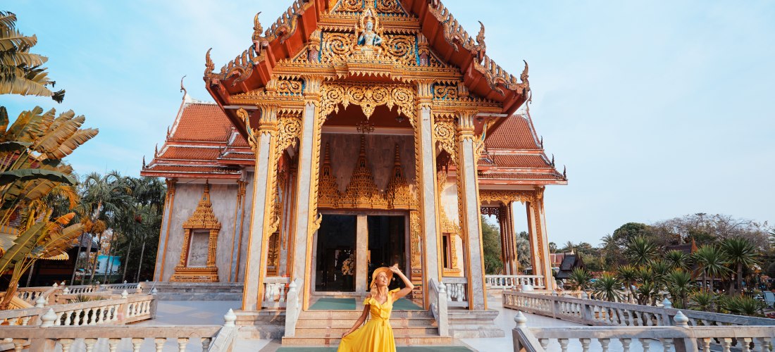 Eine Frau in einem leuchtend gelben Kleid steht auf der Treppe vor einem prachtvollen thailändischen Tempel mit kunstvoll vergoldeten Ornamenten und traditioneller Architektur