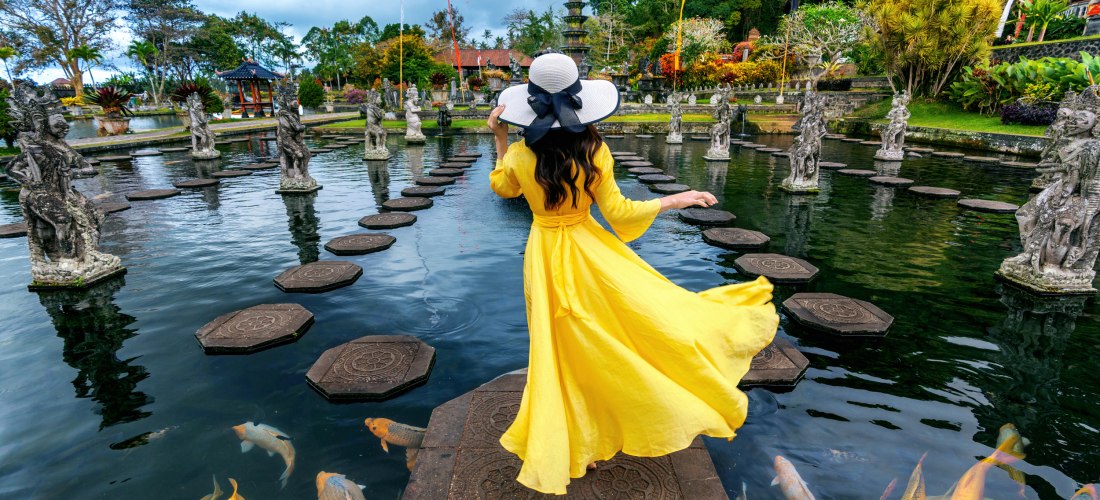 Eine Frau in einem langen, leuchtend gelben Kleid und großem Sonnenhut schreitet über Steine durch ein Wasserbecken im Tirta Gangga Wasserpalast auf Bali. Um sie herum schwimmen farbenfrohe Kois und stehen kunstvolle Steinfiguren. Der Garten ist von tropi