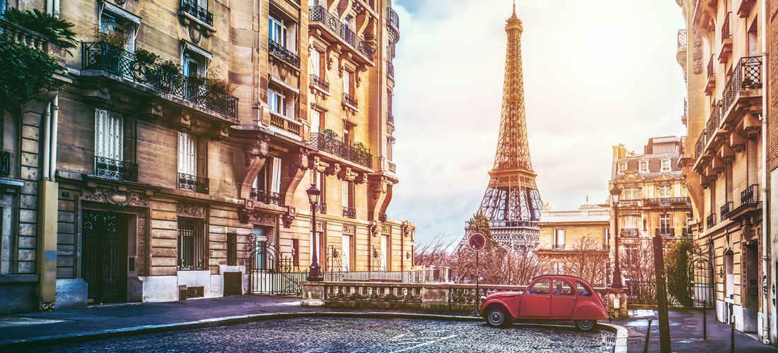 Straßenszene in Paris bei Sonnenaufgang. Links elegante Altbaufassaden mit Balkonen, rechts der Eiffelturm in voller Höhe. Im Vordergrund ein roter Oldtimer auf Kopfsteinpflaster. Weiches Licht verleiht der Szene eine romantische Stimmung.