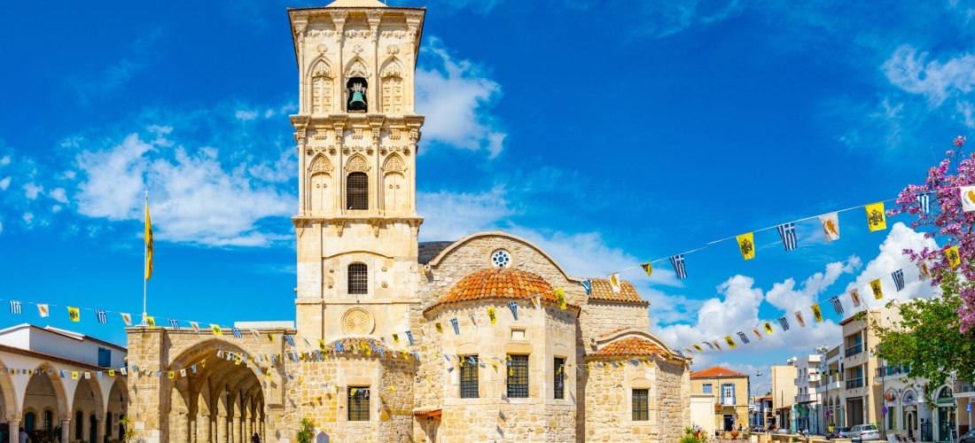 Kirche des Heiligen Lazarus in Larnaca: helles Sandsteinbauwerk mit markantem Glockenturm, Arkadengang und Wimpelketten auf dem Vorplatz.