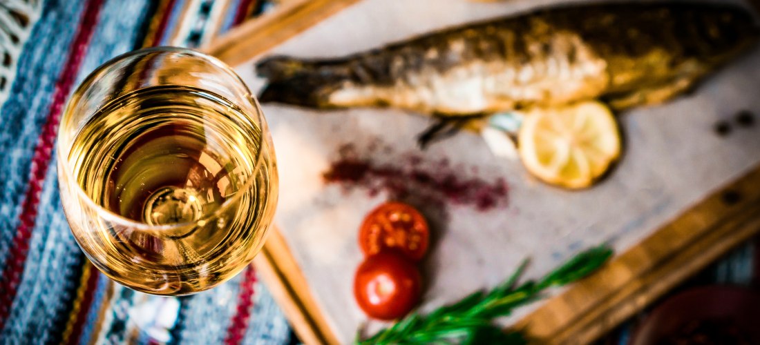 Ein gedeckter Tisch mit einem Glas Weißwein im Vordergrund und einem gegrillten ganzen Fisch im Hintergrund, serviert auf einem Holzbrett mit Zitronenscheiben, Tomaten, Rosmarin und Gemüse. Der Tisch ist mit einem bunten, gewebten Tuch bedeckt, die Atmosp