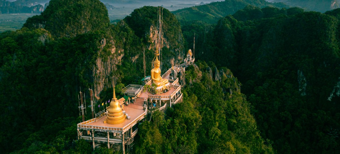 Tiger Cave Temple in Krabi, Thailand: Luftaufnahme des Tempels mit goldener Buddha-Statue auf Berggipfel inmitten tropischer Landschaft.