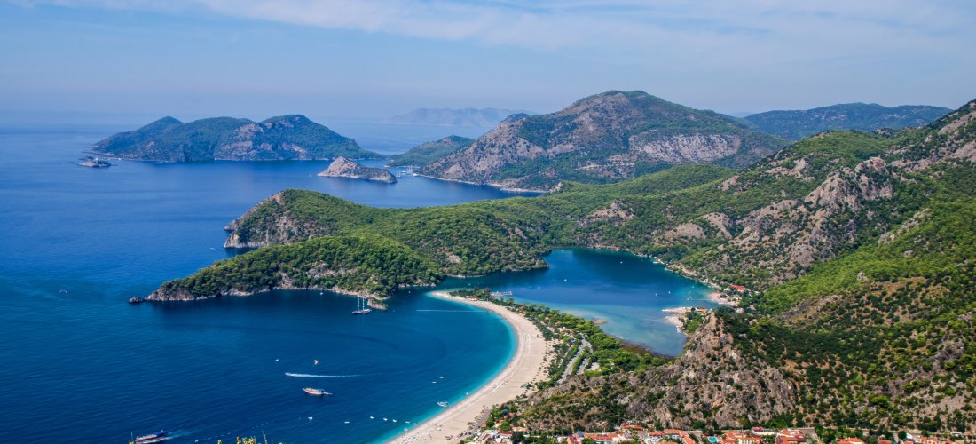 Luftaufnahme der Küstenlandschaft von Ölüdeniz an der Türkischen Riviera mit einer geschwungenen Sandbank, türkisblauem Meer, grünen Bergen und vorgelagerten Inseln.