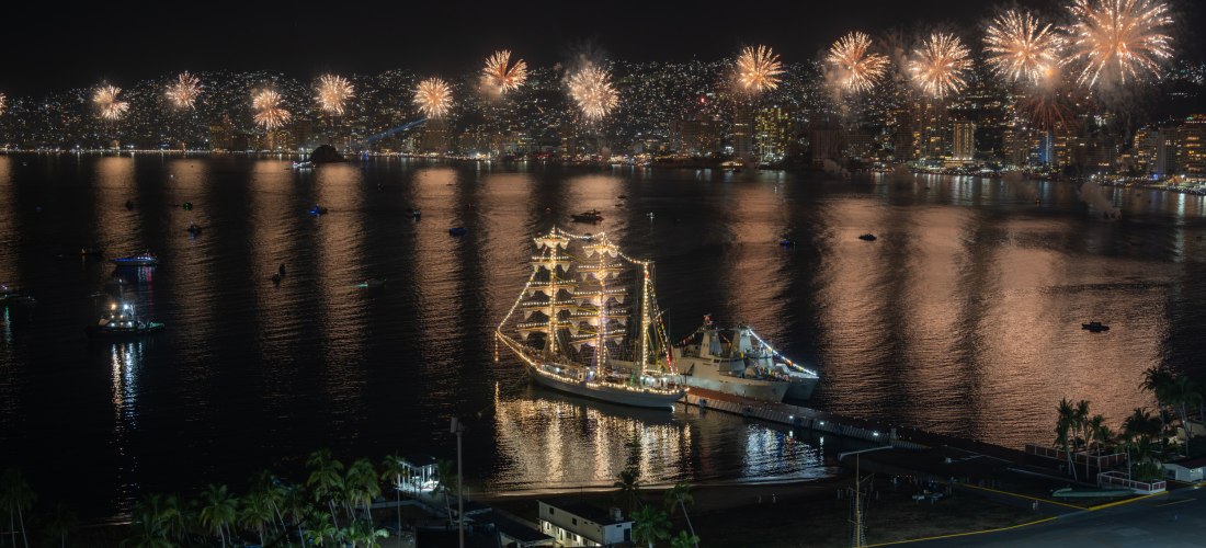 Panorama-Nachtaufnahme der Bucht von Acapulco mit spektakulärem Feuerwerk am Himmel und einem festlich beleuchteten Segelschiff im Hafen