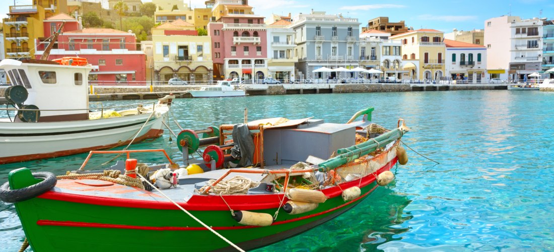 Ein traditionelles buntes Fischerboot mit grünem und rotem Rumpf schwimmt im klaren, türkisfarbenen Wasser eines Hafens auf Kreta. Im Hintergrund eine Uferpromenade mit farbenfrohen Häusern im venezianischen Stil, Cafés und Restaurants unter blauem Himmel