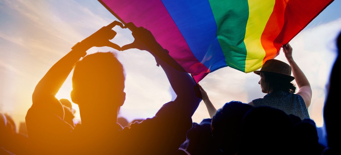 Menschen stehen in einer Menge bei Sonnenuntergang und halten eine große Regenbogenflagge über ihren Köpfen. Eine Person formt mit den Händen ein Herzsymbol. Die Szene ist in warmes Licht getaucht