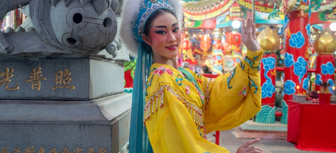Frau in farbenfrohem, gelbem traditionellen Kostüm mit Kopfschmuck. Sie posiert elegant vor einem reich geschmückten chinesischen Tempel mit roten und blauen Säulen.
