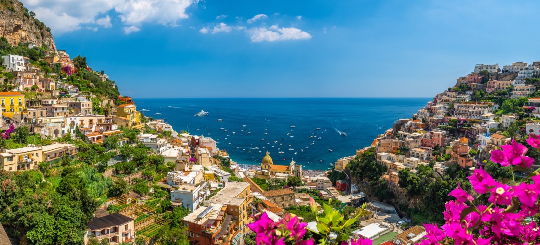 Bunte Küstenstadt an einer steilen Felsküste: terrassenförmig gebaute Häuser mit roten Dächern fallen zum tiefblauen Meer ab. Viele kleine Boote liegen in der Bucht. Im Vordergrund leuchten pinke Bougainvillea-Blüten, darüber sonniger Himmel mit wenigen W