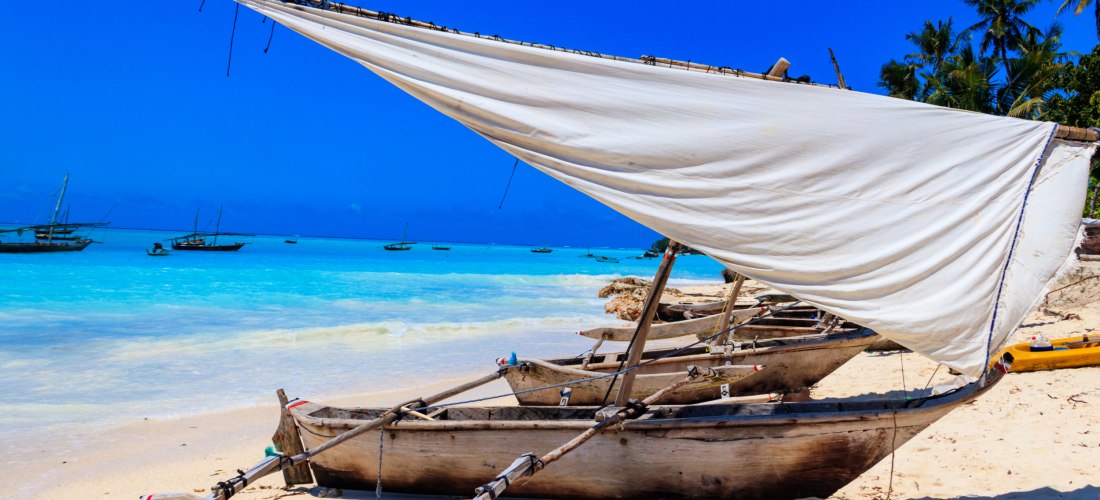 Traditionelle Holzdhaus mit weißem Lateinsegel am hellen Sandstrand; im Hintergrund türkisblaues, klares Meer und weitere Boote unter tiefblauem Himmel.
