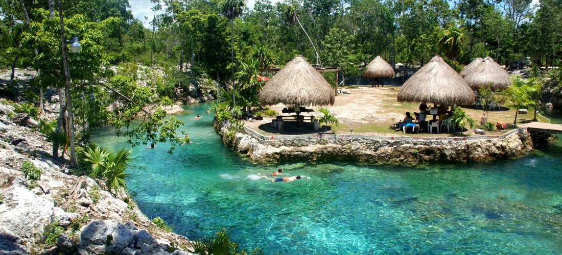 Ein paradiesischer Ort mit türkisfarbenem, kristallklarem Wasser, umgeben von üppiger tropischer Vegetation. Strohhütten bieten Schatten für Besucher, während einige Menschen das erfrischende Wasser genießen.