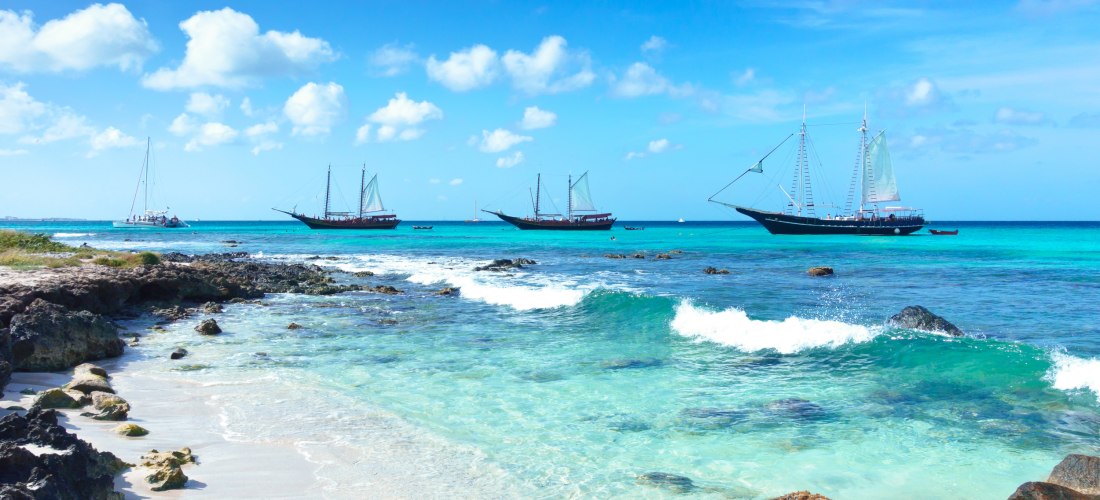 Türkisblaues Meer mit mehreren großen Segelschiffen, die vor der Küste von Aruba liegen. Im Vordergrund ein felsiger, weißer Sandstrand mit sanften Wellen und kristallklarem Wasser unter einem strahlend blauen Himmel.
