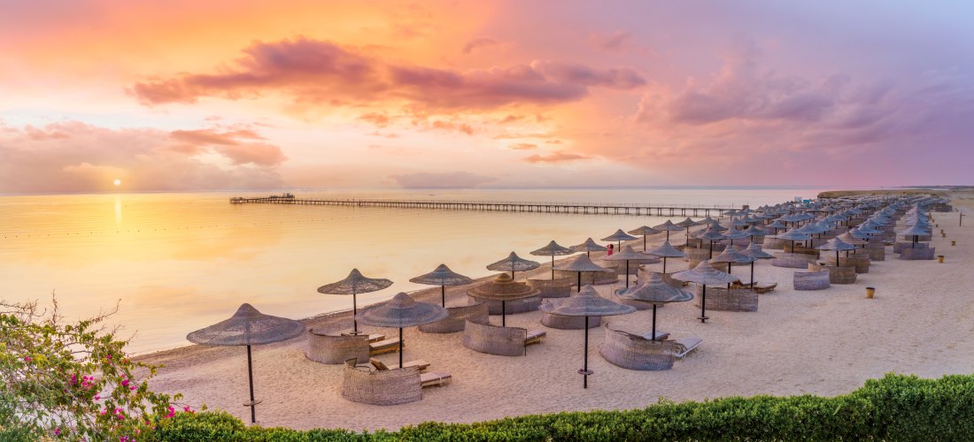 Strand bei Sonnenuntergang mit Bast-Sonnenschirmen und Liegen. Die Sonne spiegelt sich im ruhigen Meer, der Himmel ist rosa-orange. Ein langer Steg führt ins Wasser, vorne blühen pinke Blumen.