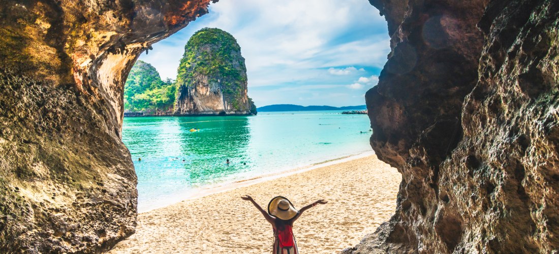Phra Nang Cave Beach in Krabi, Thailand: Frau mit Hut blickt vom Höhleneingang auf weißen Sandstrand, türkisfarbenes Meer und Karstfelsen.