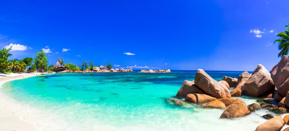 Dieses Bild zeigt eine tropische Strandlandschaft mit kristallklarem Wasser, weichem Sand und majestätischen Granitfelsen. Das ruhige, flache Wasser lädt zum Schwimmen und Entspannen ein, während die üppigen Palmen und die natürliche Felsformation eine id