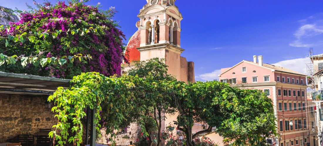 Romantisches Straßencafé in der Altstadt von Korfu mit Blick auf eine rot-goldene Kirche, umgeben von Bougainvillea und mediterranem Flair