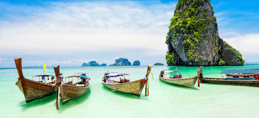 Mehrere Longtail-Boote liegen im flachen, smaragdgrünen Wasser vor einem weißen Sandstrand. Im Hintergrund ragt eine steile, grüne Kalksteininsel aus dem Meer, typisch für die Landschaft um Phuket.