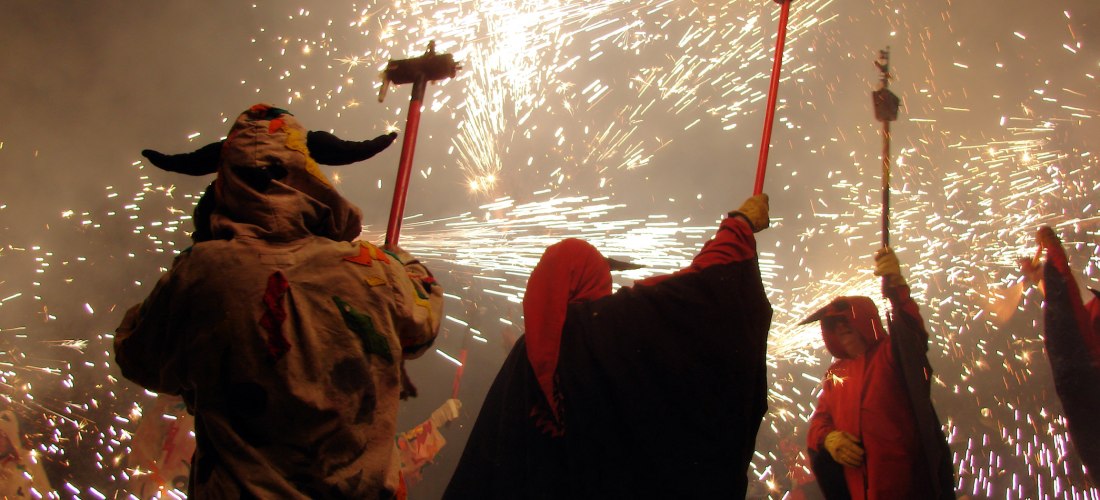 Menschen in traditionellen Kostümen mit Kapuzen und Masken schwenken Funken sprühende Feuerwerksstäbe bei Nacht. Umgeben von Rauch und Lichtblitzen während eines Festumzugs