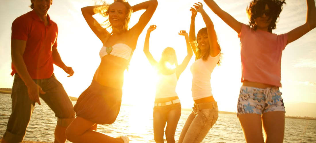 Fünf junge Menschen tanzen und springen ausgelassen am Strand im Sonnenuntergang. Die Sonne scheint direkt von hinten und wirft goldene Lichtstrahlen