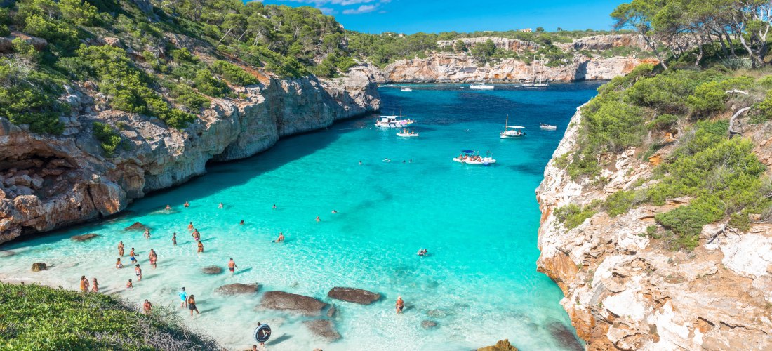 Strand Cala des Moro auf Mallorca mit kristallklarem, türkisfarbenem Wasser, Badenden im flachen Wasser und Booten in der Bucht, umgeben von Felsen und grüner Vegetation
