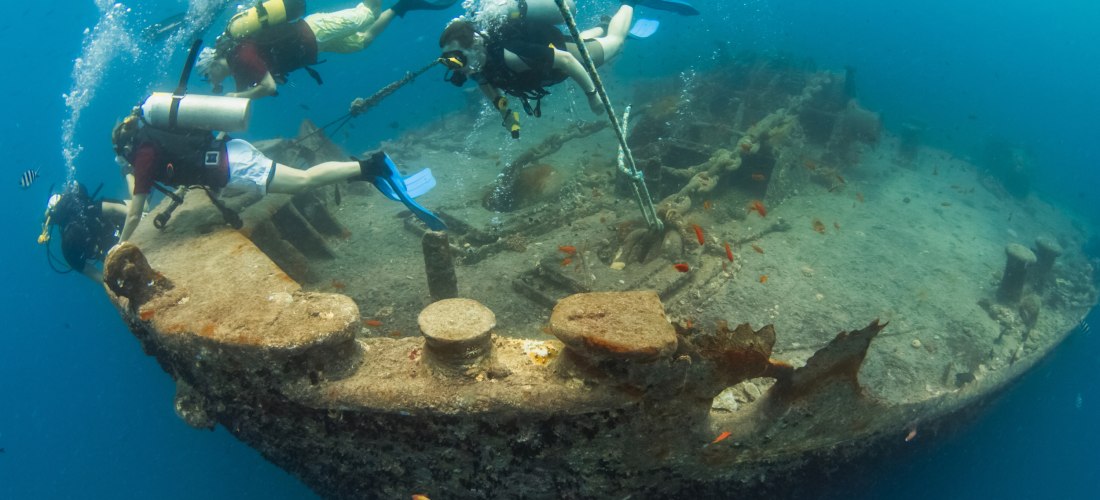 Mehrere Taucher mit Sauerstoffflaschen erforschen ein altes, rostiges Schiffswrack unter Wasser. Um sie herum schwimmen kleine bunte Fische.