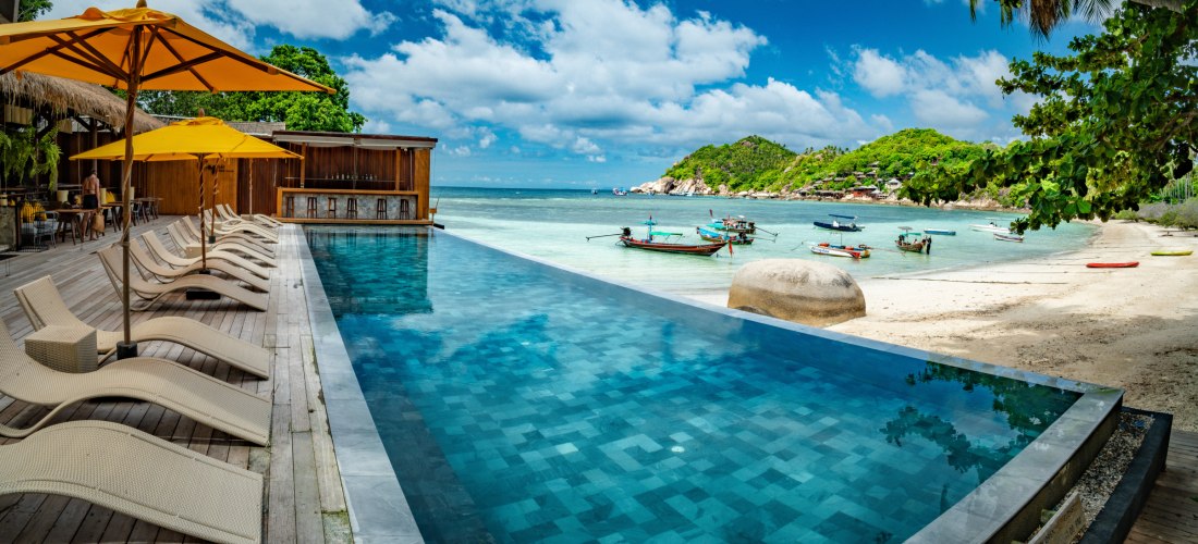Infinity-Pool mit Sonnenliegen und gelben Sonnenschirmen direkt am Strand. Im Hintergrund ruhiges Meer mit Longtail-Booten und grüne Hügel auf einer tropischen Insel.