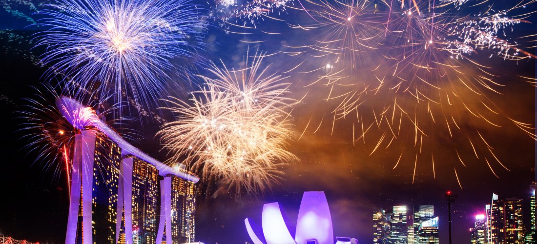 Farbenfrohes Feuerwerk über Marina Bay Sands und dem ArtScience Museum in Singapur. Die markanten Gebäude sind hell beleuchtet und spiegeln sich im Wasser der Bucht.