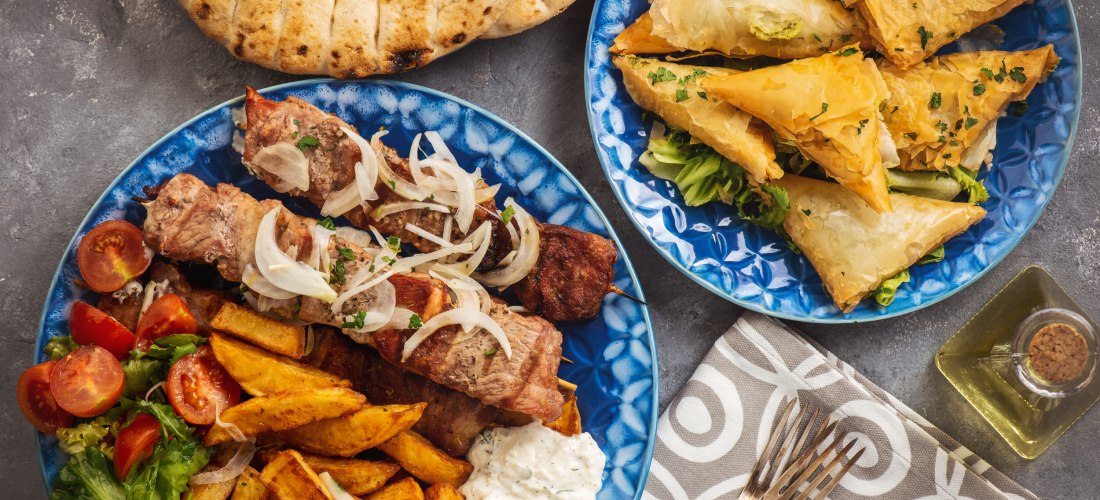 Teller mit griechischen Spezialitäten: gegrillte Fleischspieße mit Zwiebeln, Pommes frites, Tomatensalat und Tsatsiki auf einem blauen Teller. Daneben ein weiterer Teller mit goldbraun gebackenen Blätterteig-Dreiecken. Im Hintergrund Fladenbrot und eine F