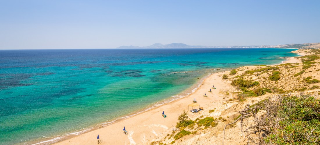 Weitläufiger, heller Sandstrand mit türkisfarbenem Wasser an der Küste einer Insel. Der Strand ist nur vereinzelt mit Liegen und Sonnenschirmen besetzt und wirkt ruhig und naturbelassen. Im Hintergrund erstreckt sich das tiefblaue Meer bis zum Horizont, w