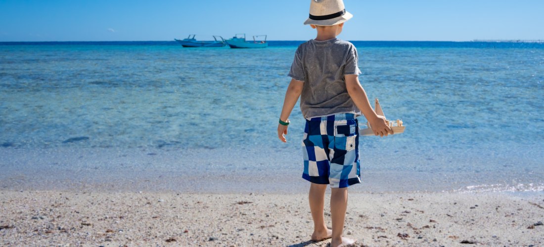 Ein kleiner Junge steht am Strand und blickt aufs Meer. Er trägt Strohhut, T-Shirt und Badeshorts und hält ein Spielzeugboot in der Hand. Im Hintergrund sind zwei Boote auf dem Wasser zu sehen.