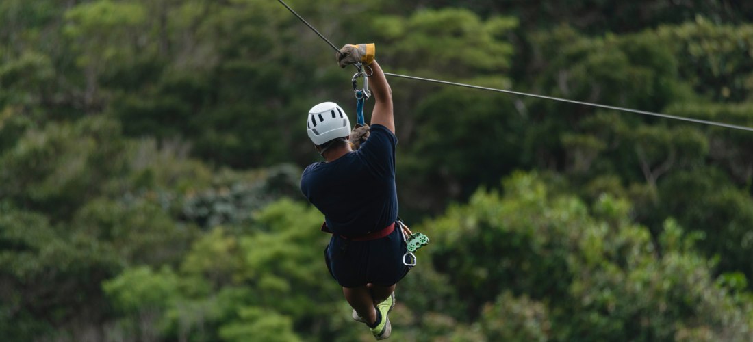 Person mit Helm beim Ziplining über einem tropischen Dschungel, Blick von hinten auf die Seilrutsche