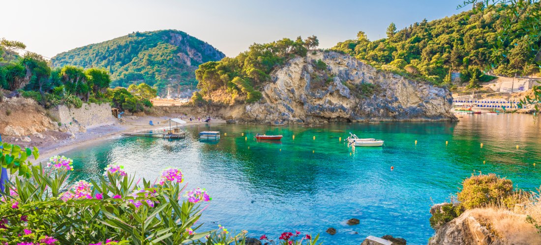 Ruhige Badebucht auf Korfu: Das glasklare Wasser leuchtet in Türkistönen, kleine Boote schaukeln sanft vor einer felsigen Halbinsel. Der Strand ist von üppigem Grün und mediterranen Blumen eingerahmt. Im Vordergrund blühen farbenprächtige Oleandersträuche