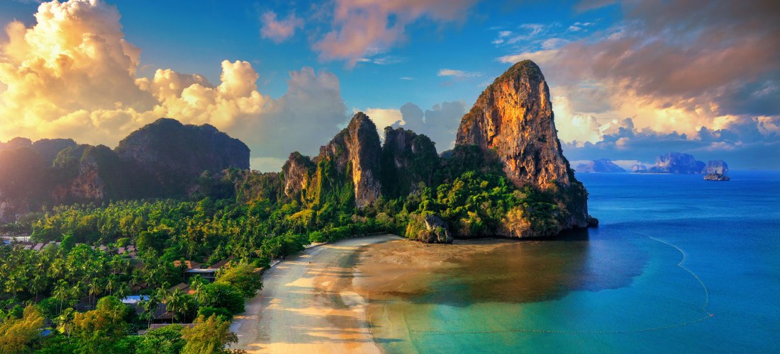 Panoramablick auf Railay Beach in Krabi bei Sonnenuntergang: goldene Wolken, türkisblaue Bucht, Longtail-Boot und steil aufragende Kalksteinfelsen an der Andamanensee.