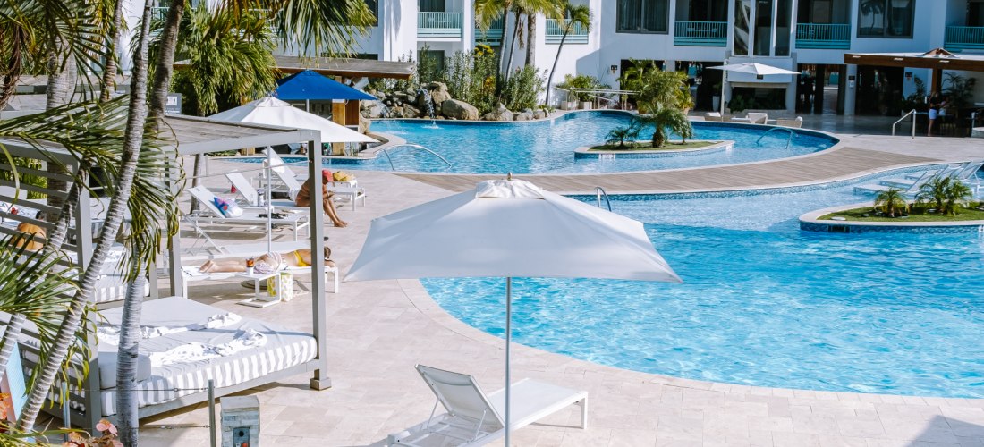 Tropische Hotelanlage auf Aruba mit weißer Fassade, Palmen und mehreren Pools. Im Vordergrund ein kleiner Whirlpool, in dem ein Paar Cocktails genießt. Im Hintergrund stehen Sonnenliegen, Sonnenschirme und weiße Gebäude mit Giebel-Dächern unter blauem Him