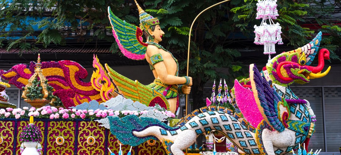 Festwagen mit detailreicher Skulptur eines geflügelten, mythologischen Wesens und einer traditionellen Figur mit Speer. Alles ist aus bunten Blüten und Perlen gestaltet, mit kunstvollen Mustern und intensiven Farben.