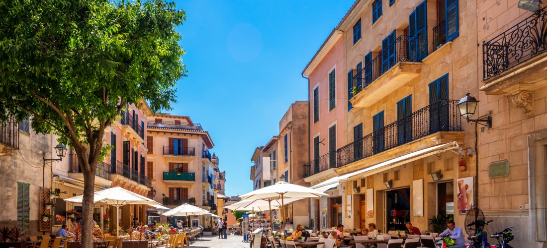Lebendiges Straßencafé in einer malerischen Altstadt auf Mallorca, mit alten Gebäuden, bunten Fensterläden, Sonnenschirmen und Menschen, die das mediterrane Flair genießen