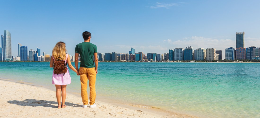Ein Paar steht am Strand und blickt auf die Skyline von Abu Dhabi mit hohen modernen Gebäuden
